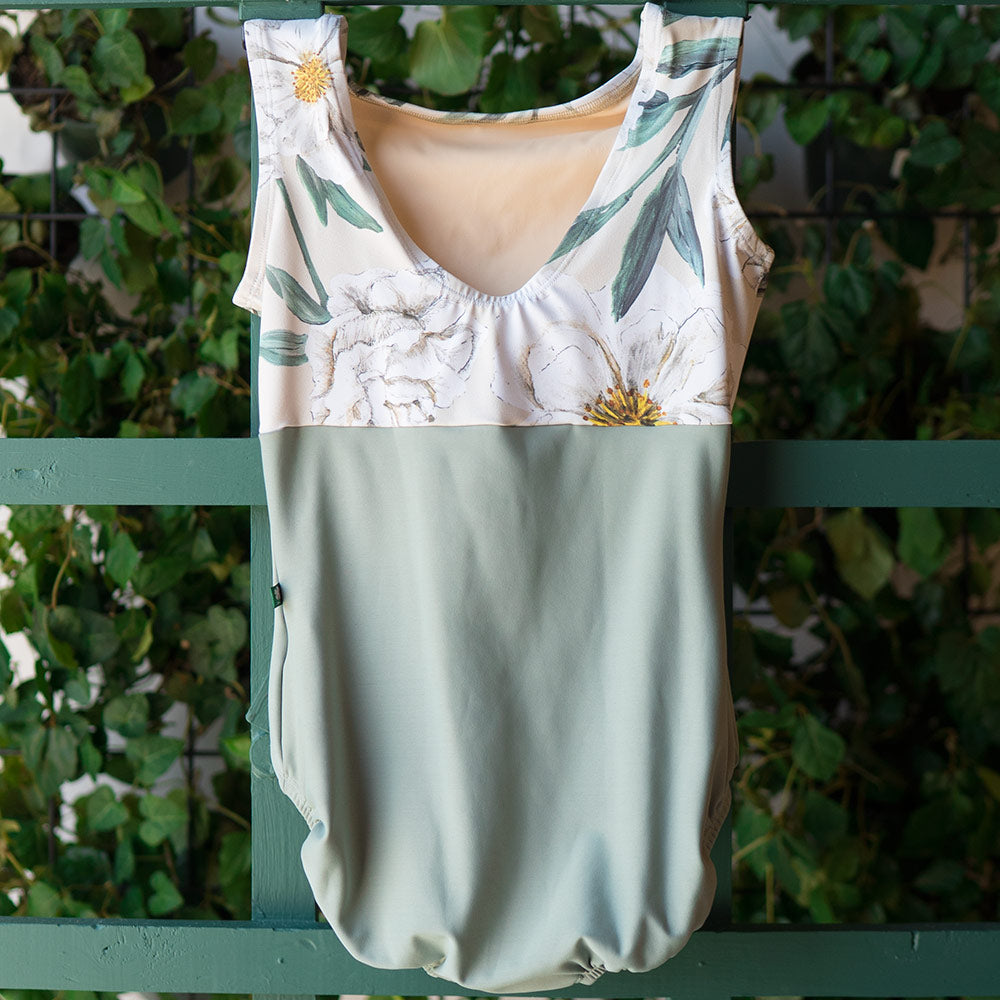 Back view of sleeveless, mid cut back ballet leotard. Bottom base in a soft, muted green aloe color and top half in an almond camellia flower print with hues of yellow, white and green